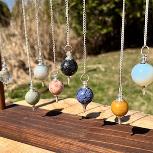 May include: A display of crystal pendulums in a range of colours and textures, hanging from silver chains. The pendulums are presented on a wooden stand, with a natural outdoor backdrop. The pendulums include clear, white, green, pink, black, blue, yellow, and opalescent spheres.
