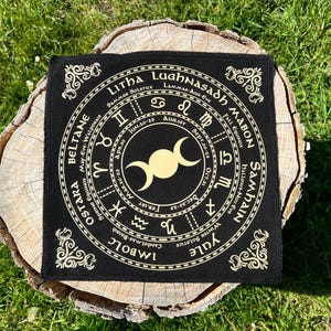May include: A black square altar cloth with gold-colored text and symbols. The cloth features a circular design with astrological symbols, seasonal names, and the phases of the moon. The cloth is placed on a tree stump.
