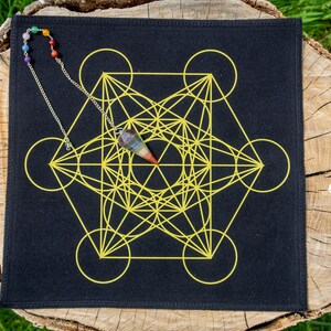 Puede incluir: Un paño cuadrado negro con un diseño amarillo del Cubo de Metatrón. Un péndulo con un cristal de arcoíris y una cadena de cuentas cuelga sobre el paño. Los artículos se exhiben sobre una superficie de madera con hierba verde al fondo.