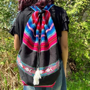 May include: A black backpack with a colorful striped design in red, blue, and pink. The backpack has a drawstring closure and a white tassel. The straps are also striped. The backpack is worn over the shoulders.