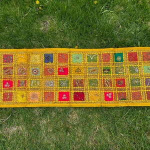 Op de afbeelding: Een rechthoekig patchworktextiel met een gele rand en een raster van kleurrijke vierkanten. De vierkanten hebben verschillende patronen en kleuren, waaronder rood, groen, blauw en goud. Het textiel ligt op groen gras.