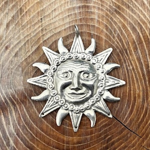May include: A silver-colored sun pendant with a smiling face, surrounded by pointed rays and a beaded border. The pendant is displayed on a wooden surface with visible grain patterns.