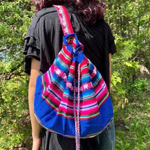 Peut inclure: Un sac à dos tissé coloré avec une fermeture à cordon. Le sac présente un motif vibrant de rayures bleues, roses, rouges et blanches, avec une base bleue unie. La bandoulière est rouge avec des accents floraux blancs.