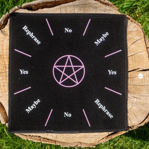 Puede incluir: Una esterilla de adivinación negra con un pentagrama rosa en el centro y las palabras "Yes", "No", "Maybe" y "Rephrase" en los bordes. La esterilla está colocada sobre un tocón de madera con hierba verde de fondo.