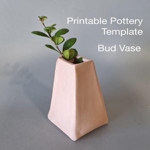 Könnte beinhalten: Druckbare Töpfer-Vorlage für eine Knospenvase, eine rosafarbene Keramikvase mit quadratischem Boden und dreieckiger Form. Eine kleine grüne Pflanze steht in der Vase.