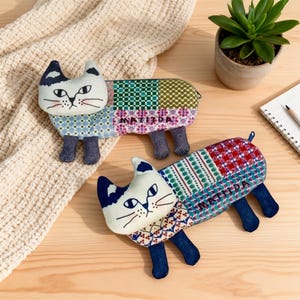 Puede incluir: Dos monederos de tela hechos a mano con forma de gato y diseños de patchwork. Los gatos tienen caras blancas con detalles en negro y azul, y la palabra "MATILDA" está bordada en el cuerpo. Un gato está sobre una manta de punto color crema, y el otro sobre una superficie de madera.