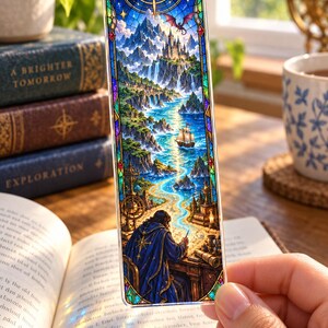 May include: A rectangular bookmark featuring a stained-glass style illustration of a person in a blue cloak writing at a desk. The scene depicts a winding river, a sailing ship, a castle on a mountain, a dragon, and a crescent moon in a blue sky.