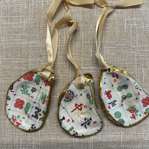 May include: Three oyster shell ornaments with gold trim and attached gold ribbon. Each shell is decorated with a Mahjong tile design in red, green, and black. The shells are on a textured beige surface.
