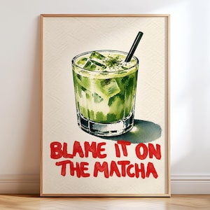 May include: A framed wall art print featuring a watercolour illustration of a glass filled with a green matcha beverage, ice cubes, and a black straw. Below the glass, the text BLAME IT ON THE MATCHA is written in bold red capital letters.