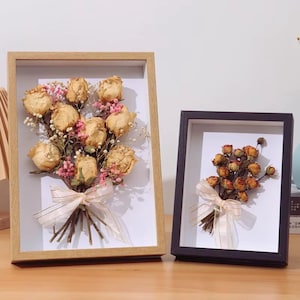May include: Two framed floral arrangements. One features a bouquet of dried roses in a light wood frame, tied with a cream ribbon. The other has a similar arrangement in a black frame. Both are on a light brown surface.
