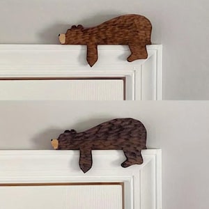 Può includere: Due statuette di orso in legno, una marrone chiaro e una marrone scuro, sono appoggiate su un telaio di porta bianco. Gli orsi sono scolpiti con dettagli strutturati e i loro musi sono di una tonalità più chiara di marrone. Gli orsi sono decorativi.