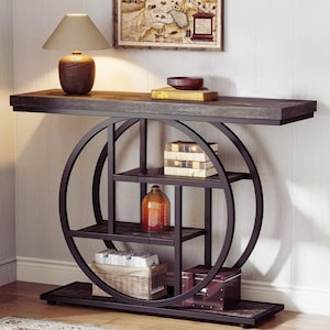 May include: A console table featuring a dark wood rectangular top and a black metal frame with a large circular design. The structure includes multiple interior shelves holding books, a glass bottle, a woven basket, and a small storage box.