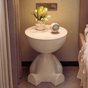 Può includere: Un tavolino bianco e moderno dal design unico. Il tavolo presenta un piano rotondo, una sezione centrale sferica e una base a forma di figura seduta. Un piccolo vaso bianco con fiori gialli e una piccola sveglia sono appoggiati sopra. Il tavolo è alto circa 61 cm.