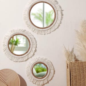May include: Three round wall mirrors in a boho style. Each mirror has a circular frame wrapped in brown rope, surrounded by a white fringe. The mirrors are mounted on a white wall, creating a decorative display.