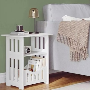 Puede incluir: Una mesa auxiliar de madera blanca con diseño de listones, con dos estantes y un revistero. Una lámpara pequeña, libros y una cámara están sobre la mesa. Un sofá gris y una manta con flecos están en el fondo.