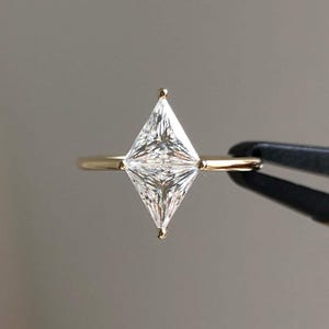 May include: A gold ring featuring a single kite-shaped diamond set in a four-prong setting. The diamond is clear and faceted, displaying a sharp, elongated geometric silhouette. The band is a smooth, polished gold metal, held by black tweezers.