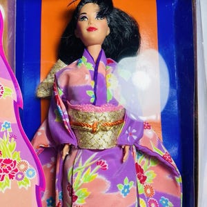 Puede incluir: Muñeca con un kimono japonés tradicional, con un diseño floral morado y rosa. La muñeca tiene cabello negro y lleva un obi dorado. El kimono presenta un estampado floral con detalles verdes, rojos y blancos. La muñeca está en una caja con fondo azul y naranja.
