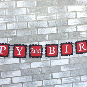May include: A red and black banner with the words "HAPPY 2nd BIRTHDAY" in white letters. The banner has a scalloped edge and features a Minnie Mouse silhouette on each end. The banner is suspended against a grey brick wall.