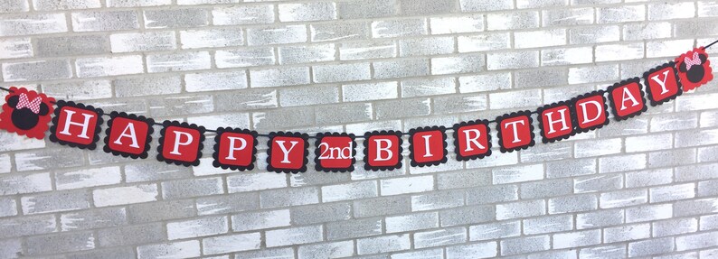 May include: A red and black banner with the words "HAPPY 2nd BIRTHDAY" in white letters. The banner has a scalloped edge and features a Minnie Mouse silhouette on each end. The banner is suspended against a grey brick wall.