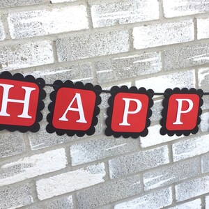 May include: A red and black banner with the words "HAPPY 2nd B" in white letters. The banner has a scalloped edge and a Minnie Mouse head on the left side. The banner is hanging in front of a grey brick wall.
