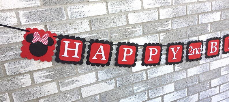 May include: A red and black banner with the words "HAPPY 2nd B" in white letters. The banner has a scalloped edge and a Minnie Mouse head on the left side. The banner is hanging in front of a grey brick wall.