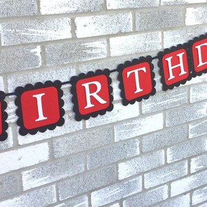 May include: A red and black birthday banner with white lettering spelling out "BIRTHDAY". The banner is strung on a black string and is hanging against a white brick wall. The banner has a scalloped edge.