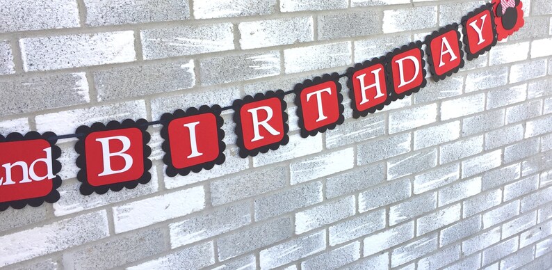 May include: A red and black birthday banner with white lettering spelling out "BIRTHDAY". The banner is strung on a black string and is hanging against a white brick wall. The banner has a scalloped edge.