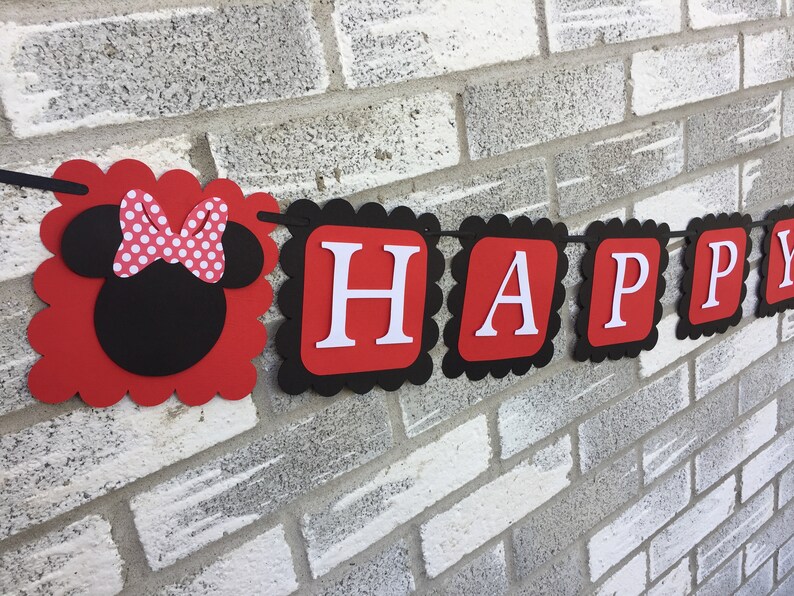 May include: A red and black banner spelling "HAPPY" in white letters. The banner features a Minnie Mouse head with a polka dot bow. The banner has a scalloped edge and is strung on a black string, suitable for a party.