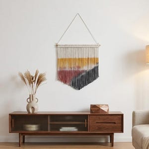 May include: A wall hanging made of off-white cotton cord featuring a dip-dyed design with horizontal bands of mustard yellow, muted rose pink, and charcoal gray. The fiber art piece is suspended from a wooden dowel by a triangular cord loop.