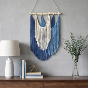 May include: A wall hanging featuring layered macrame cords in shades of deep navy, light blue, and cream. The cords are suspended from a horizontal wooden branch by a white string, creating a tiered, curved fringe design with a textured appearance.