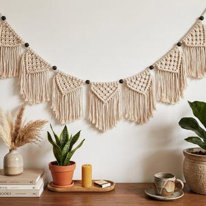 May include: A macrame wall hanging featuring seven triangular, knotted off-white cotton cord pennants with long fringe, separated by dark wooden beads on a string. The banner is suspended horizontally against a plain wall above various home decor items.