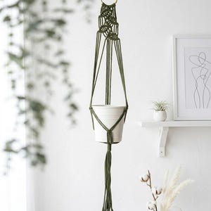 May include: A hanging olive green macrame plant holder featuring a series of intricate knotted cords that support a plain white cylindrical pot. The structure is suspended from a circular gold-toned metal ring at the top and ends in a tassel.