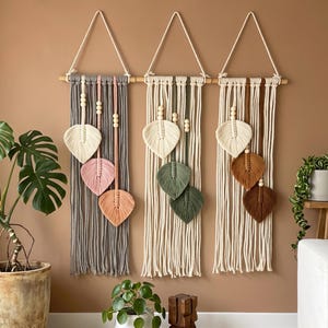 May include: Three macrame wall hangings suspended from wooden dowels with white rope loops. Each piece features vertical cord strands, wooden beads, and three leaf-shaped fiber elements in varying shades of gray, pink, peach, cream, green, and brown.