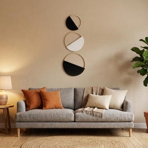 May include: A living room setting with a grey sofa, orange and cream cushions, a wooden side table with a lamp, and a potted plant. The wall features three circular decorative pieces with black, white, and wood finishes.