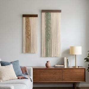May include: Two macrame wall hangings with wooden tops. One is beige with a brown leaf design, the other is beige with a green leaf design. They hang on a light gray wall above a wooden cabinet.