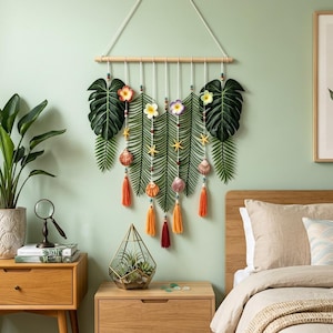 May include: A wall hanging featuring a wooden dowel with hanging white cords, adorned with green monstera and fern leaves, colorful plumeria flowers, small yellow starfish, seashells, and dangling orange and dark red fabric tassels.
