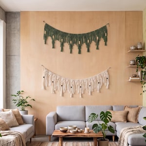 May include: Two macrame wall hangings suspended on a wooden wall. The top piece is dark green with light wooden beads, and the bottom piece is off-white with dark wooden beads. Both feature intricate knotted patterns and long hanging fringe tassels.