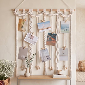 May include: A wall-hanging macrame photo display featuring off-white cotton cord knotted in various patterns, suspended from a horizontal wooden dowel. The piece includes several vertical strands with small wooden beads and wooden clips for photos.
