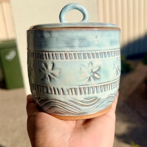 May include: A light blue ceramic jar with a matching lid featuring a circular handle. The exterior surface displays embossed floral patterns, horizontal bands of vertical dashes, and wavy lines near the base, revealing a natural clay colour.