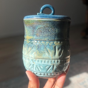 May include: A cylindrical ceramic jar with a lid featuring a small circular handle. The exterior is glazed in shades of blue and green with a textured, embossed pattern of repeating leaf-like shapes around the lower half and a smooth upper rim.