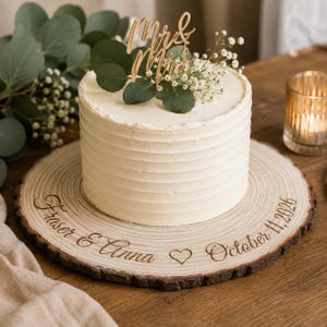 May include: A white frosted cake sits on a round wooden slice engraved with the text Fraser & Anna, a heart symbol, and October 11, 2026. The cake is topped with a wooden Mr & Mrs cake topper, green eucalyptus leaves, and white baby's breath flowers.