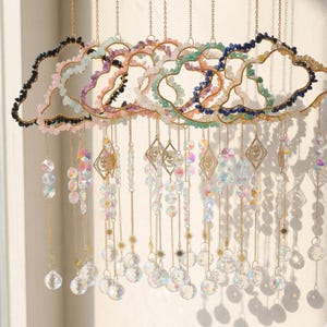 Op de afbeelding: Een decoratieve hangende mobiel met wolkvormige frames, versierd met kleurrijke edelstenen. Gouden kettingen hangen kristallen kralen, facetgeslepen prisma's en ruitvormige bedels op. Ontworpen om het licht te vangen en een sprankelend effect te creëren.