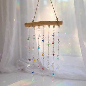 May include: A decorative wind chime featuring a natural wooden bar suspended by twine. Silver chains hang from the bar, adorned with colorful glass beads and crystal pendants. The beads are in various colors, including blue, green, red, and yellow.