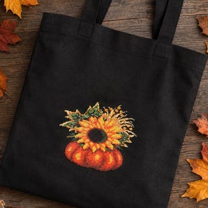 May include: A black tote bag featuring a cross-stitched design of a vibrant orange pumpkin topped with a large yellow sunflower, green leaves, and wheat stalks. The bag has two black fabric handles and a simple, rectangular construction.