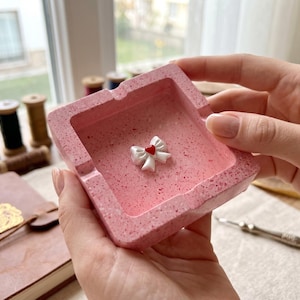 Puede incluir: Un cenicero cuadrado rosa con textura moteada, con un lazo blanco con un corazón rojo en el centro. El cenicero es sostenido por una persona. Artículo decorativo.