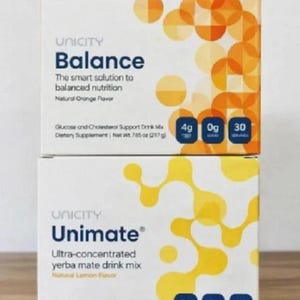 May include: Two white boxes of Unicity dietary supplements. The top box is "Balance" with "Natural Orange Flavor" and nutritional information. The bottom box is "Unimate" with "Natural Lemon Flavor" and nutritional information.