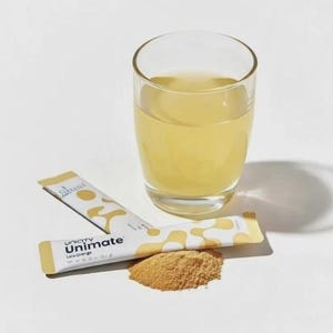 K&ouml;nnte beinhalten: Ein klares Glas gef&uuml;llt mit einer blassgelben Fl&uuml;ssigkeit, begleitet von zwei wei&szlig;en und gelben Unimate Lemon Orange Getr&auml;nkepulver-Sticks und einem kleinen Haufen hellbraunem Pulver, alles auf einer schlichten wei&szlig;en Oberfl&auml;che angeordnet.