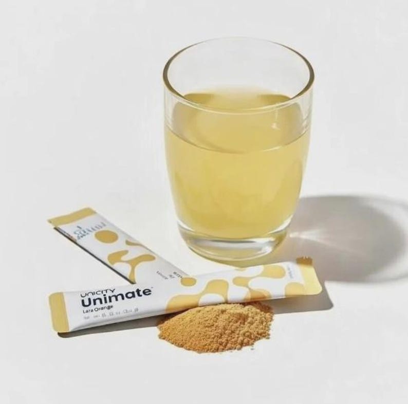 K&ouml;nnte beinhalten: Ein klares Glas gef&uuml;llt mit einer blassgelben Fl&uuml;ssigkeit, begleitet von zwei wei&szlig;en und gelben Unimate Lemon Orange Getr&auml;nkepulver-Sticks und einem kleinen Haufen hellbraunem Pulver, alles auf einer schlichten wei&szlig;en Oberfl&auml;che angeordnet.