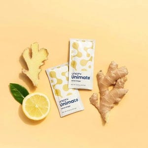 K&ouml;nnte beinhalten: Zwei rechteckige Unicity Unimate Lemon Ginger Beutel mit einem gelb-wei&szlig;en abstrakten Muster, jeweils beschriftet mit net wt. 6.3 g, begleitet von einer frischen Zitronenscheibe mit einem gr&uuml;nen Blatt und zwei St&uuml;cken Ingwerwurzel.