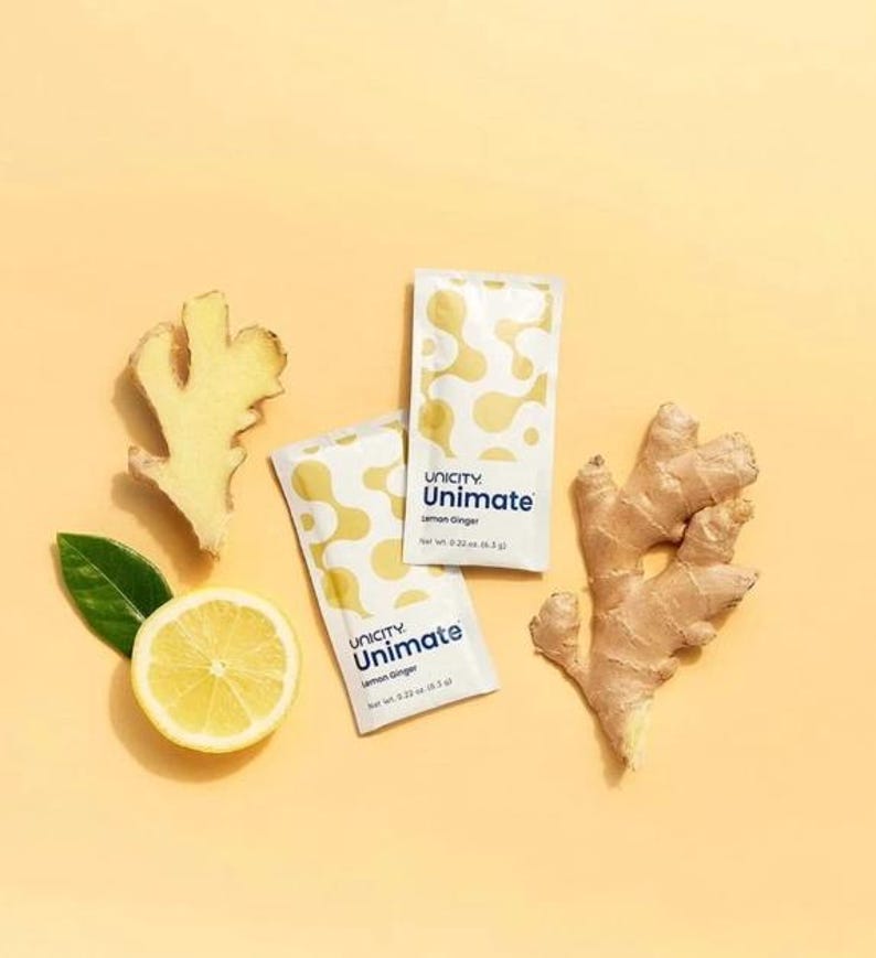 K&ouml;nnte beinhalten: Zwei rechteckige Unicity Unimate Lemon Ginger Beutel mit einem gelb-wei&szlig;en abstrakten Muster, jeweils beschriftet mit net wt. 6.3 g, begleitet von einer frischen Zitronenscheibe mit einem gr&uuml;nen Blatt und zwei St&uuml;cken Ingwerwurzel.
